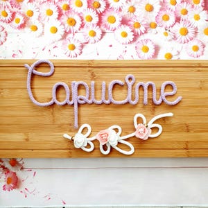 May include: A light purple knitted cord spelling the name Capucine in cursive script, mounted on a rectangular bamboo board. Below the name, a white knitted cord forms decorative loops accented with small pink and white fabric flowers.