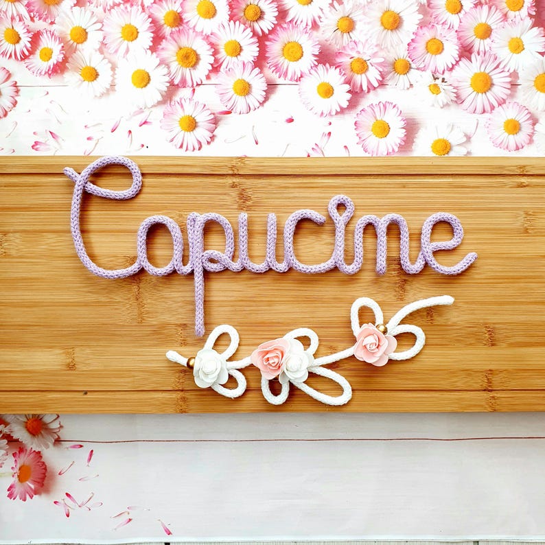 May include: A light purple knitted cord spelling the name Capucine in cursive script, mounted on a rectangular bamboo board. Below the name, a white knitted cord forms decorative loops accented with small pink and white fabric flowers.