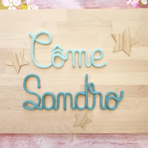 May include: A wall decoration featuring the words C&ocirc;me and Sandro formed from light blue and teal knitted yarn cord. The display includes three wooden star-shaped cutouts arranged around the cursive text on a light wood grain background.
