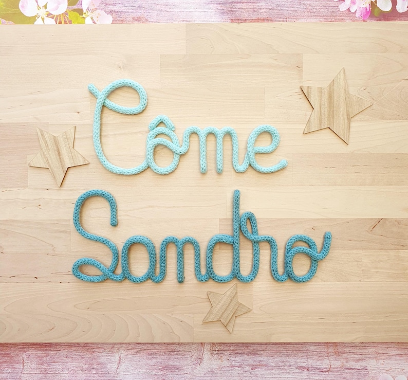May include: A wall decoration featuring the words C&ocirc;me and Sandro formed from light blue and teal knitted yarn cord. The display includes three wooden star-shaped cutouts arranged around the cursive text on a light wood grain background.