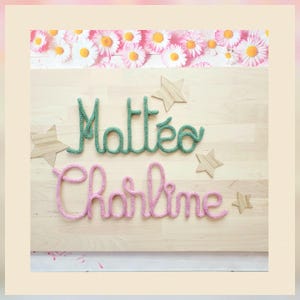 May include: A light wood board featuring the names Matteo in green yarn and Charline in pink yarn, written in a cursive script. The board is decorated with four light wood star cutouts and a border of white and yellow daisy flowers.
