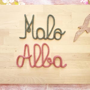 May include: Two cursive words crafted from knitted yarn. The top word reads Malo in a muted sage green color, and the bottom word reads Alba in a dusty rose pink color. A small wooden bird cutout is positioned to the right of the green word.