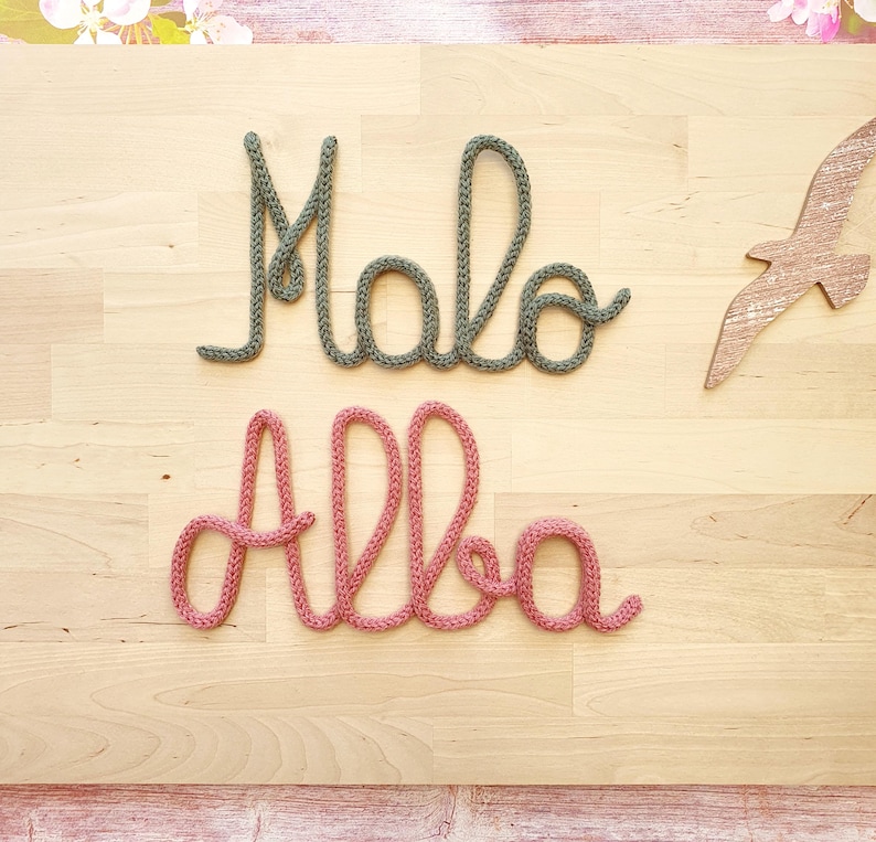 May include: Two cursive words crafted from knitted yarn. The top word reads Malo in a muted sage green color, and the bottom word reads Alba in a dusty rose pink color. A small wooden bird cutout is positioned to the right of the green word.