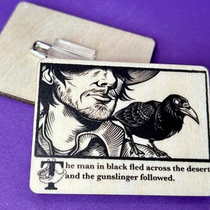 May include: Wooden pin badge with a black and white illustration of a man and a raven, and the text: "The man in black fled across the desert, and the gunslinger followed." A safety pin is attached.