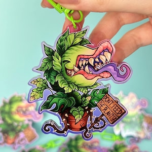 May include: A clear acrylic keychain depicting a cartoon plant with a large mouth and the text "DON'T FEED THE PLANTS!". The plant is green with pink and purple details, and is attached to a green clasp.