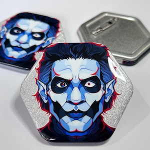 May include: Hexagonal button badges with a stylized illustration of a character with blue and white face paint, dark hair, and red accents. The badges have a silver glitter border and a pin back.