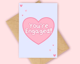 Könnte beinhalten: Eine Grußkarte mit dem Text "You're Engaged!" in einem rosa Herzen auf hellblauem Hintergrund. Die Karte liegt auf einem braunen Umschlag, mit kleinen rosa Herzen verstreut. Der Hintergrund ist einfarbig hellrosa.