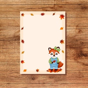 May include: A rectangular notepad with a cream background and an autumnal leaf border. A cartoon fox in blue dungarees and a green bow is on the bottom right. The notepad is set against a wooden surface.