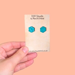 Może przedstawiać: Para turkusowych niebieskich kolczyków w kształcie kości D20 na kartce z napisem "D20 Studs by Peach & Mimi" i "www.peachandmimi.co.uk".
