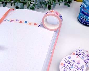 Può includere: Un rotolo di nastro washi rosa e bianco con un motivo floreale. Il nastro è largo 15 mm e lungo 10 m. Il nastro è etichettato "Flower Power Washi Tape by Peach & Mimi".