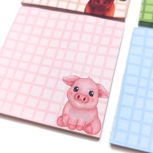May include: A stack of square notepads with a grid pattern. One notepad features a pink background with a cute cartoon pig illustration. Other notepads have beige and blue backgrounds with grid patterns.