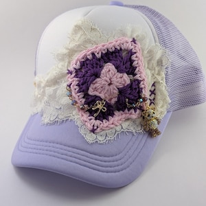 May include: A lavender baseball cap with a white front panel and a mesh back. The cap is adorned with a crocheted square in shades of purple and pink, accented with lace and a small charm of a teddy bear.