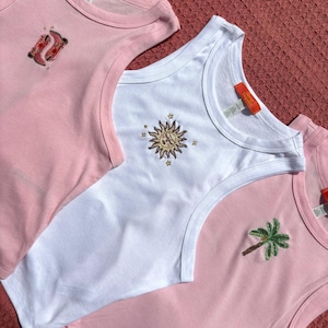 May include: Three sleeveless tank tops featuring embroidered designs. One light pink top displays red cowboy boots, a white top features a gold sun with stars, and another light pink top shows a green palm tree. Each garment has an orange neck label.