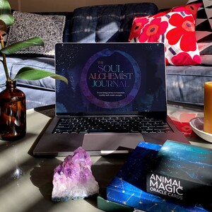 May include: A laptop computer with a screen displaying the text "The Soul Alchemist Journal: A year-long journey to transmute earthly and cosmic energies". A purple amethyst crystal sits on a table next to the laptop. A deck of oracle cards with the text "Animal Magic Oracle Cards" is on the table.