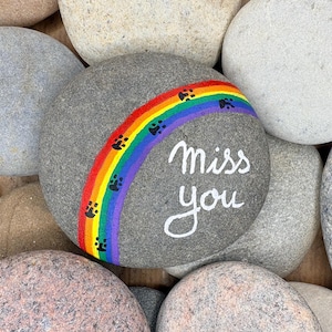 Può includere: Un sasso di fiume grigio liscio dipinto con un vivace arcobaleno che si inarca sulla superficie. Piccole impronte di zampe nere sono dipinte lungo l'arcobaleno e le parole miss you sono scritte in bianco sul lato destro della pietra.