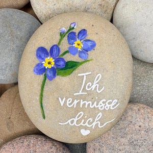 Könnte beinhalten: Ein glatter, hellbrauner Flusskiesel, handbemalt mit zwei blauen Vergissmeinnicht-Blüten mit gelben Zentren und grünen Blättern. Der weiße Text "Ich vermisse dich" ist in Schreibschrift unter den Blumen zu sehen, gefolgt von einem kleinen Herz.