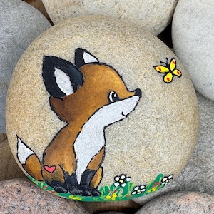 Könnte beinhalten: Ein glatter, runder, beigefarbener Stein mit einer handgemalten Illustration eines braunen Fuchses mit weißer Brust und Schwanzspitze, schwarzen Ohren und Pfoten sowie einem kleinen roten Herz an der Seite, der einen gelben Schmetterling betrachtet.