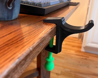 May include: A black plastic desk clamp with a green knob and a hook extending out. The clamp is attached to a wooden desk.