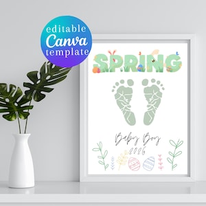 May include: A framed print with the word "SPRING" in green, decorated with Easter-themed elements. Below are green footprints and the text "Baby Boy 2026". The print is in a white frame, with a white vase and green plant to the left.