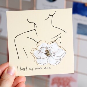 以下が含まれることがあります： ベージュのカードに、黒い線で描かれた人物が白い花を胸に抱えているイラストが描かれています。カードの下部には黒い文字で「I trust my inner voice.」と書かれています。