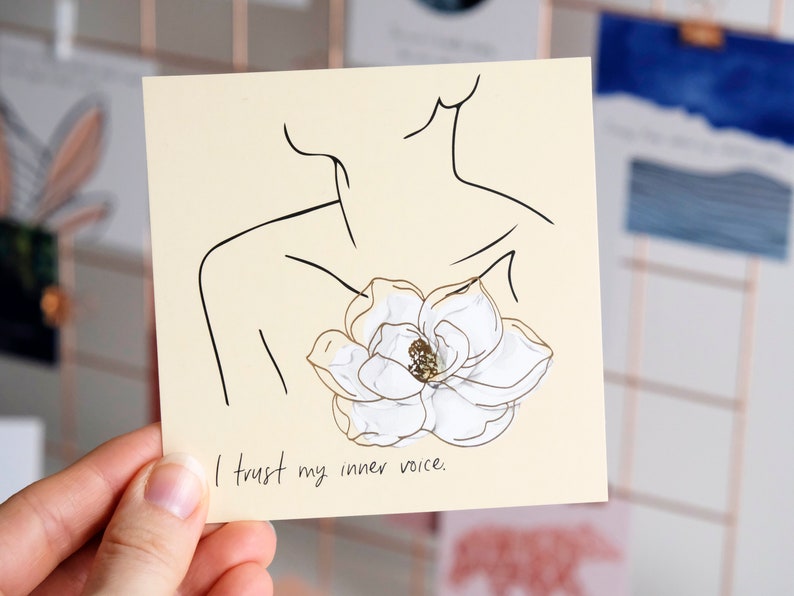 以下が含まれることがあります： ベージュのカードに、黒い線で描かれた人物が白い花を胸に抱えているイラストが描かれています。カードの下部には黒い文字で「I trust my inner voice.」と書かれています。