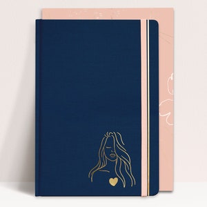 May include: A navy blue notebook with a gold outline of a woman with a heart on her chest. The notebook has a pink elastic band closure and a pink and white patterned cover.