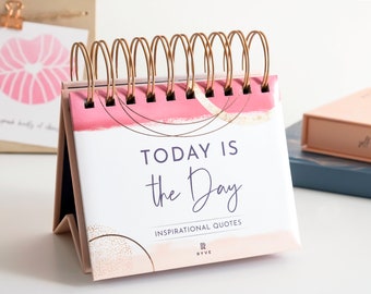 May include: A pink and gold desktop calendar with a white background. The calendar features a gold spiral binding and the text "TODAY IS the Day" in purple lettering. The calendar also includes the text "INSPIRATIONAL QUOTES" and the logo "RYVE".