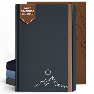 May include: A dark blue Daily Gratitude Journal with a mountain and sun graphic. The journal has a brown elastic band and a brown tag that reads "DAILY GRATITUDE JOURNAL". The journal is next to a brown journal with a mountain graphic.