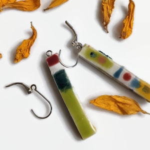 May include: A pair of colorful ceramic earrings with silver hooks. The earrings are rectangular and feature abstract patterns in shades of green, yellow, red, and blue.