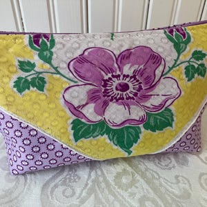 May include: A rectangular cosmetic bag with a purple zipper. The bag features a large purple flower with green leaves on a white background, framed by yellow and purple patterned fabric. The bag is approximately 8 inches wide.