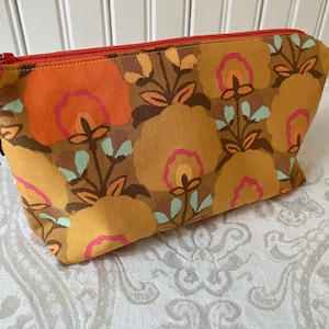May include: A rectangular cosmetic bag with a floral pattern in shades of orange, yellow, brown, and pink. The bag has a red zipper and a tassel with a turquoise bead. The bag is likely made of fabric.