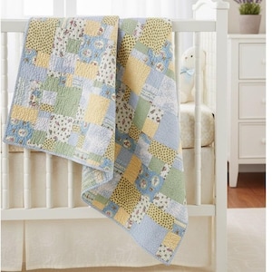 May include: A patchwork baby quilt in shades of blue, yellow, and green, draped over a white crib. The quilt features a variety of floral and geometric patterns, creating a cozy and inviting aesthetic. The quilt is approximately 40 inches by 45 inches.