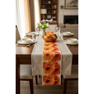 May include: A dining table set for a meal, featuring a beige table runner with a repeating orange floral pattern. The table is set with white plates, bowls, and gold-colored cutlery. Clear wine glasses and a wooden bowl of fruit complete the setting.