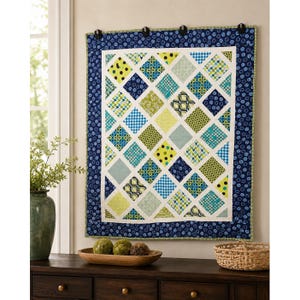 May include: A rectangular wall quilt featuring a diamond-patterned center with various shades of blue, green, and white fabric squares. The quilt is framed by a dark blue border with a circular pattern and is hung by four black clips.