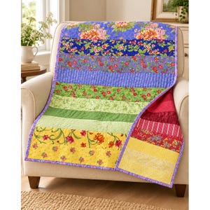 May include: A patchwork quilt featuring horizontal stripes of various floral and patterned fabrics in shades of blue, green, yellow, and red. The quilt has a purple border and is draped over a beige upholstered armchair.