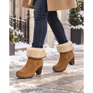 May include: Tan suede ankle boots with a thick, cream-colored shearling cuff, a block heel, and a row of small metal studs along the base. The boots are worn with dark blue denim jeans and a tan coat, standing on a snowy outdoor path.