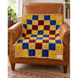 May include: A patchwork quilt featuring a repeating grid of yellow, blue, and maroon fabric squares. The yellow squares have a small multi-colored pattern, while the blue and maroon squares are solid. A white label reads "Quilts by Annie Books by Amanda".