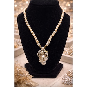 May include: A cream pearl necklace on a black display stand. The necklace has a pendant of large, round pearls, embellished with clear crystals and smaller pearls. A classic piece of jewellery.