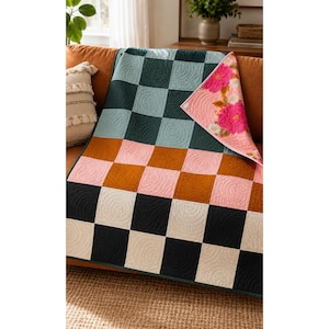 May include: A quilted throw blanket featuring a checkered pattern in shades of dark teal, light blue, rust orange, soft pink, cream, and black. The underside of the folded corner reveals a floral print with bright pink and white flowers on a pink background.