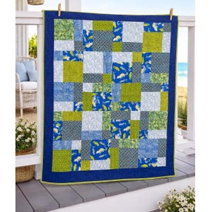 May include: A patchwork quilt with a blue border and a green inner border. The quilt features squares of various blue, green, and white fabrics. Some squares have dinosaur patterns. The quilt is hanging on a white railing.