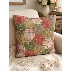 May include: A square throw pillow featuring a patchwork design of hexagonal fabric pieces. The quilt-style cover displays a variety of floral, leaf, and plaid patterns in shades of sage green, dusty rose, cream, and muted red, with visible stitching.