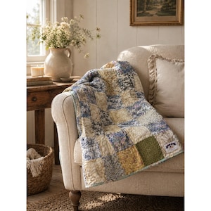 May include: A patchwork quilt featuring a variety of square fabric blocks in shades of blue, cream, yellow, and green. The quilt has a visible stitched texture and a small rectangular white label with dark text sewn onto the bottom right corner.