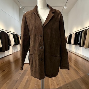 May include: A brown suede blazer with a notched collar, two front pockets, and three buttons. The jacket is displayed on a mannequin in a well-lit room with clothing racks in the background.