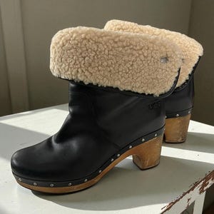 May include: Black leather ankle boots featuring a fold-over cuff lined with cream-colored shearling. The boots have a wooden block heel and a wooden sole base accented with silver-toned metal studs along the perimeter. The UGG logo is embossed.