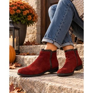May include: A pair of deep red suede ankle boots featuring a black trim along the side with small metallic studs, a side zipper closure, and a low heel. The boots are worn with blue denim jeans, showcasing a rounded toe and a sleek, flat profile.