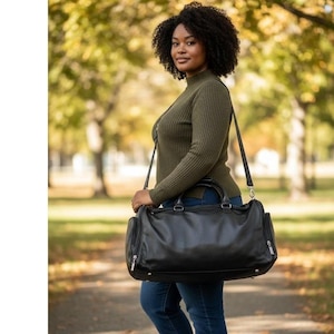 May include: A black leather duffel bag with silver-toned hardware. The bag has a shoulder strap and two handles. The bag has two zippered side pockets. The person is wearing a green sweater and blue jeans.