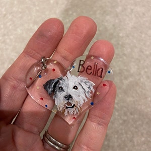 May include: Clear heart-shaped pet tag with a painted portrait of a white and gray dog. The name "Bella" is written in red script. The tag has a silver metal ring for attachment and is decorated with red, blue, and yellow dots.