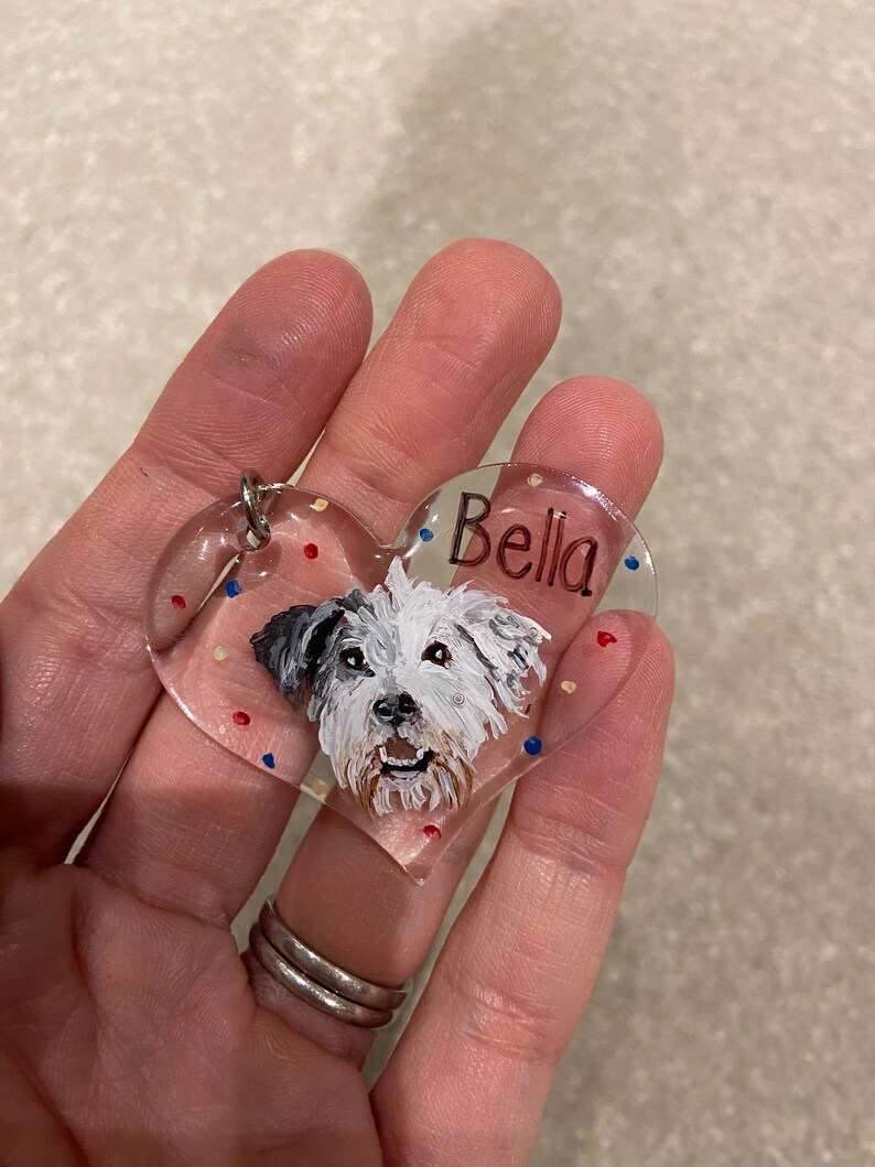 May include: Clear heart-shaped pet tag with a painted portrait of a white and gray dog. The name "Bella" is written in red script. The tag has a silver metal ring for attachment and is decorated with red, blue, and yellow dots.