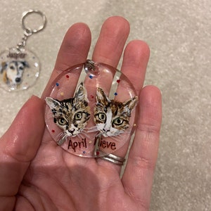 May include: A clear, round keychain featuring two cat portraits with the names "April" and "Nieve." The cats have brown and white fur. The keychain has a silver-colored ring and chain. A second keychain with a dog portrait is visible in the background.
