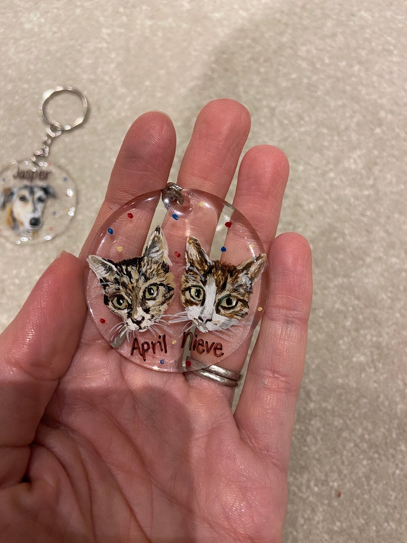 May include: A clear, round keychain featuring two cat portraits with the names "April" and "Nieve." The cats have brown and white fur. The keychain has a silver-colored ring and chain. A second keychain with a dog portrait is visible in the background.